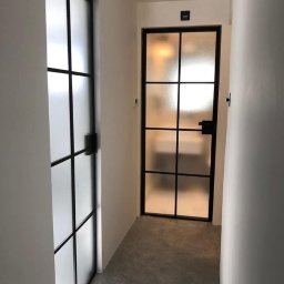 Metalovo - Dwie czarne, metalowe drzwi wewnętrzne z matowym szkłem w wąskim korytarzu z szarą wykładziną. Drzwi po lewej stronie są przesuwne.