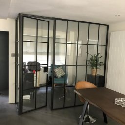 Metalovo - Loftowa ścianka działowa z czarnymi szprosami i otwartymi drzwiami, oddzielająca przestrzeń biurową w Sanoku. Widoczny stół i krzesło.