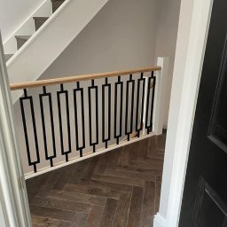 Metalovo - Nowoczesna balustrada schodowa z czarnymi, geometrycznymi elementami i drewnianym poręczą, na tle parkietu ułożonego w jodełkę.