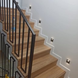 Metalovo - Drewniane schody z czarną, metalową balustradą i oświetleniem punktowym na ścianie. Widoczna butelka wody na jednym ze stopni. Ujęcie z dołu.