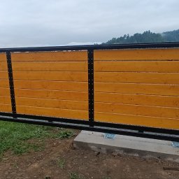 Metalovo - Nowoczesna brama przesuwna z drewnianym wypełnieniem i czarną, metalową ramą na tle pagórkowatego krajobrazu. Widoczne detale montażowe.