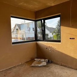 Oleksandr Petruk - Wnętrze po nałożeniu tynku maszynowego, widoczne okno narożne z czarną ramą, fragment instalacji elektrycznej i hydraulicznej, oraz worki z zaprawą na podłodze.