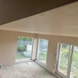 Wnętrze po nałożeniu gładzi gipsowej na ściany i sufit, widoczne okna z widokiem na zewnątrz, na podłodze resztki materiałów budowlanych, w rogu stoi mężczyzna w zielonej koszulce.