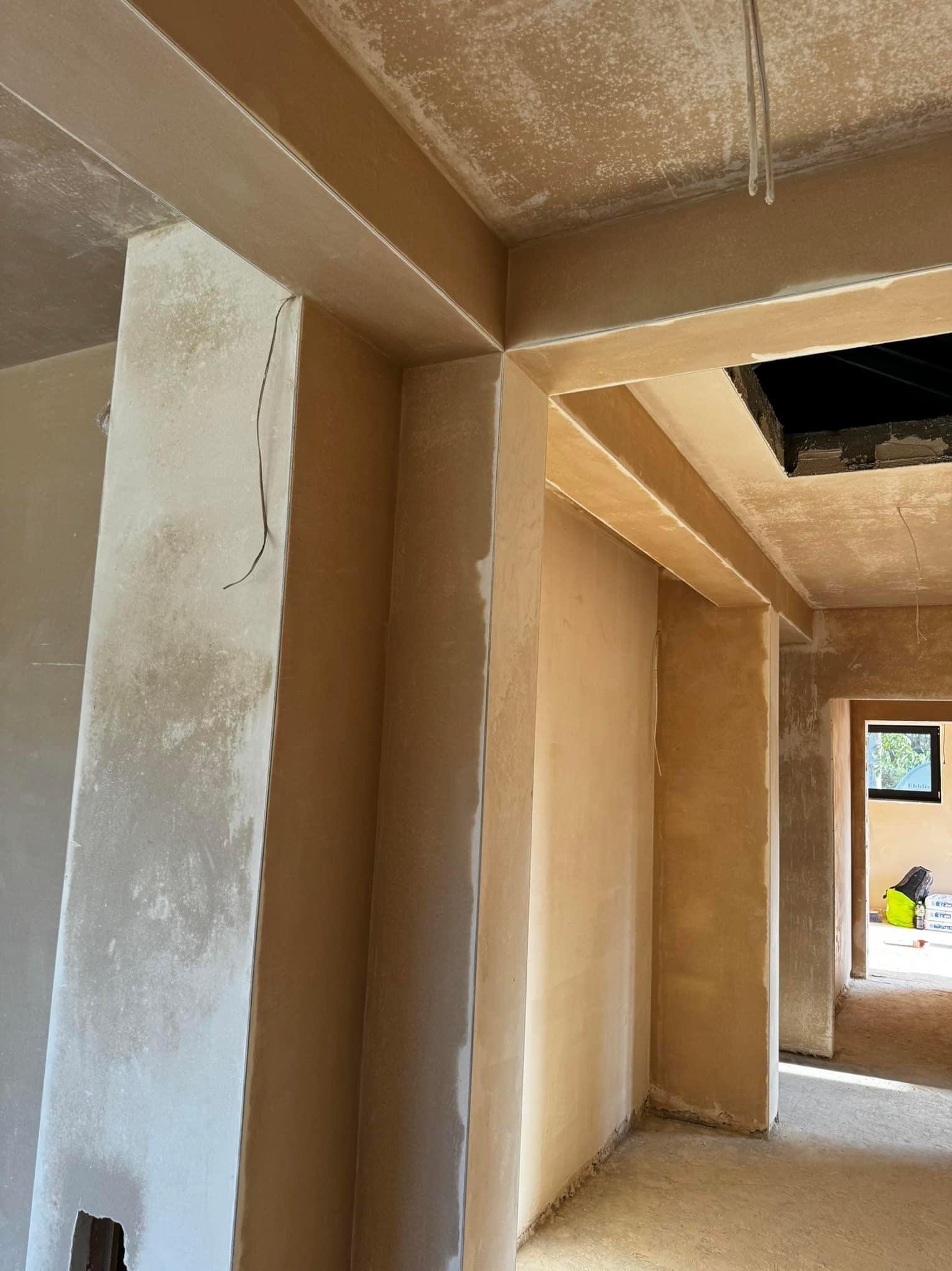 Świeżo nałożone tynki maszynowe na ścianach i sufitach w surowym wnętrzu, widoczne nierówności i zacieki, w tle okno i worki z zaprawą.