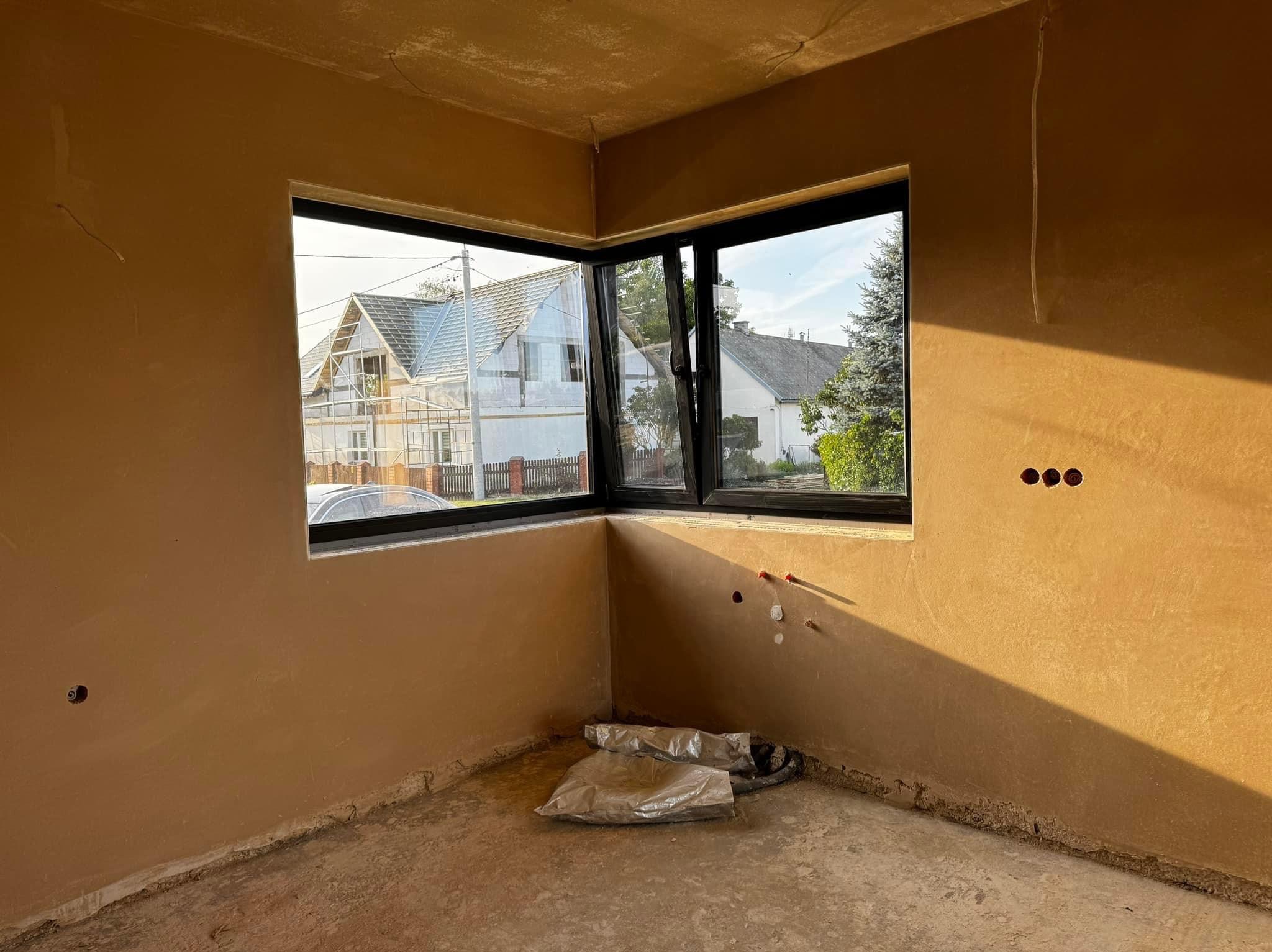 Wnętrze po nałożeniu tynku maszynowego, widoczne okno narożne z czarną ramą, fragment instalacji elektrycznej i hydraulicznej, oraz worki z zaprawą na podłodze.