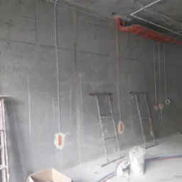 Surowy betonowy pokój w trakcie instalacji elektrycznej: widoczne kable prowadzone po ścianach i suficie, pomarańczowe rury, zaznaczone miejsca na puszki elektryczne, metalowe drabiny, karton...