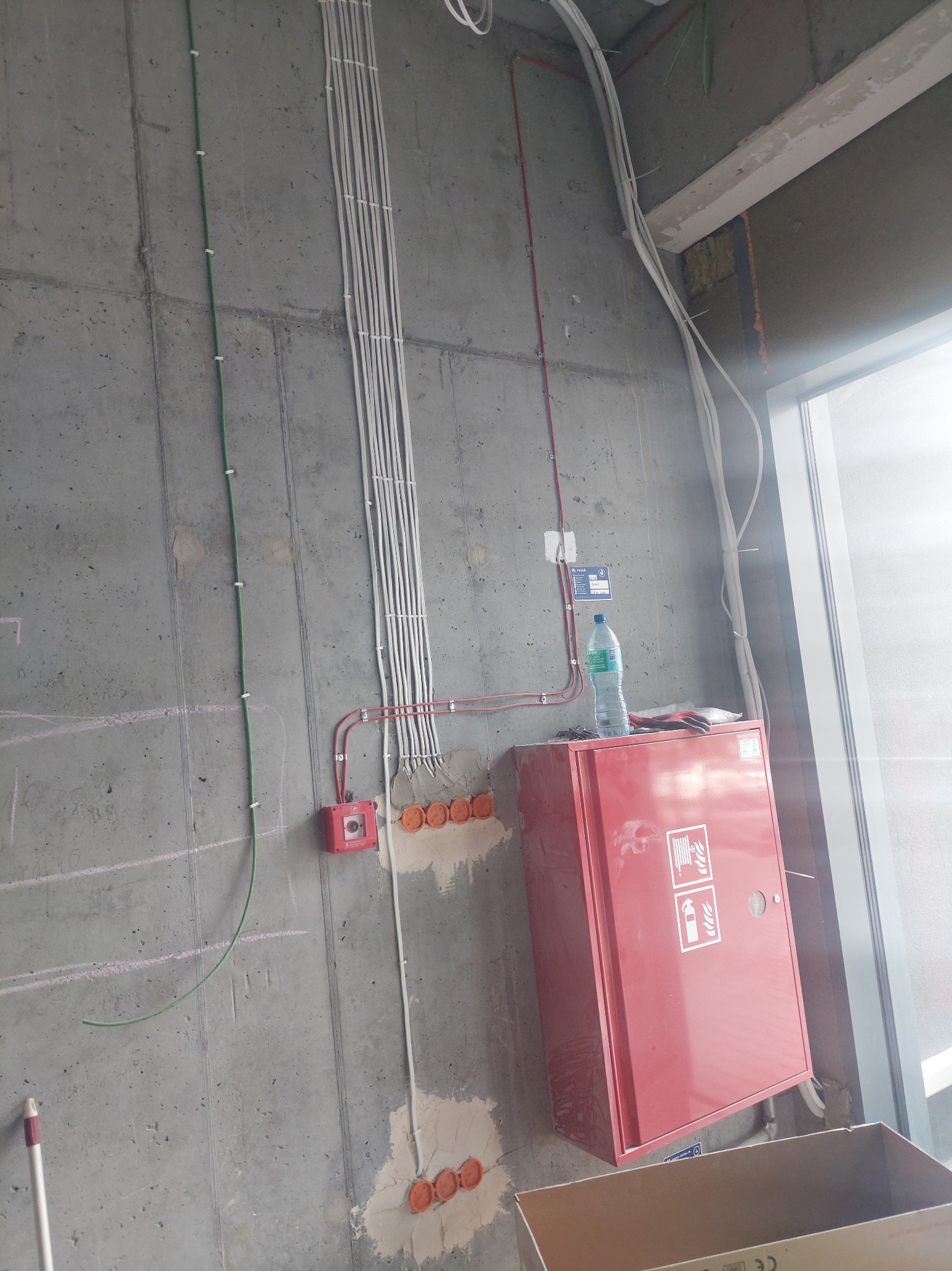 Surowa ściana z widocznymi przewodami elektrycznymi w białych listwach, puszkami instalacyjnymi i czerwoną skrzynką hydrantową, butelka wody i rękawice na skrzynce, etap prac wykończeniowych.