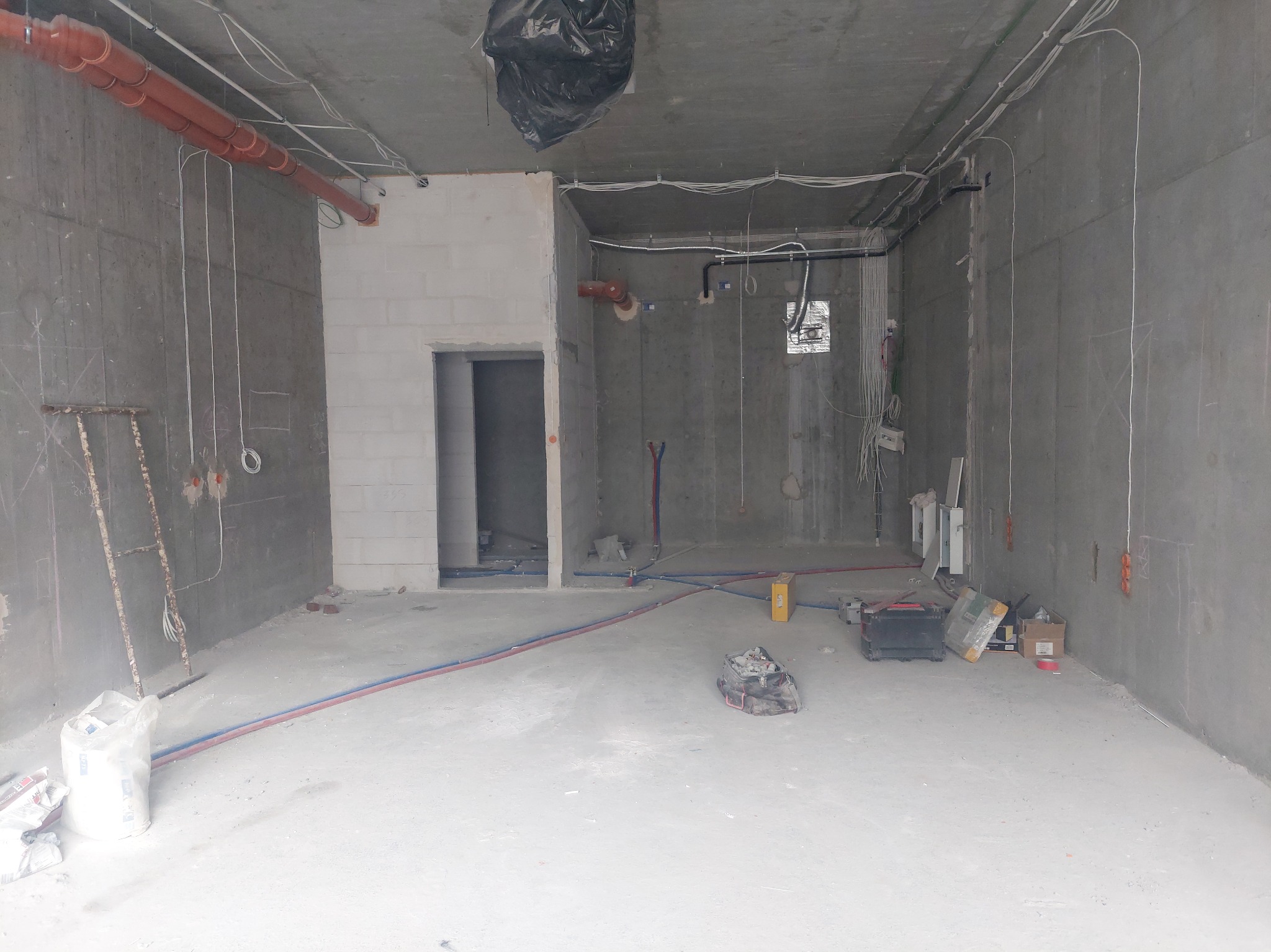 Surowy stan wnętrza w trakcie prac instalacyjnych: widoczne przewody elektryczne pod sufitem, rury wentylacyjne, instalacja wodna, ściany w betonie, narzędzia i materiały budowlane na podłodze.