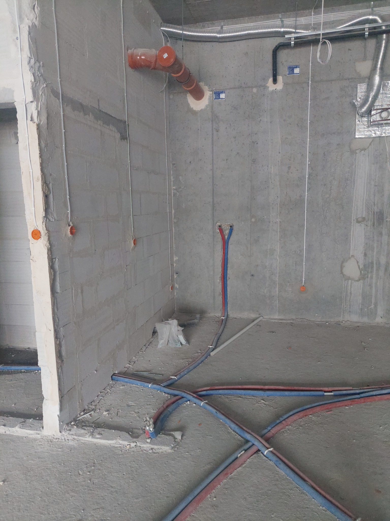 Surowy betonowy pokój w trakcie prac wykończeniowych z widocznymi kablami elektrycznymi, rurami wentylacyjnymi i instalacją wodną w kolorach niebieskim i czerwonym, wychodzącymi ze ściany.