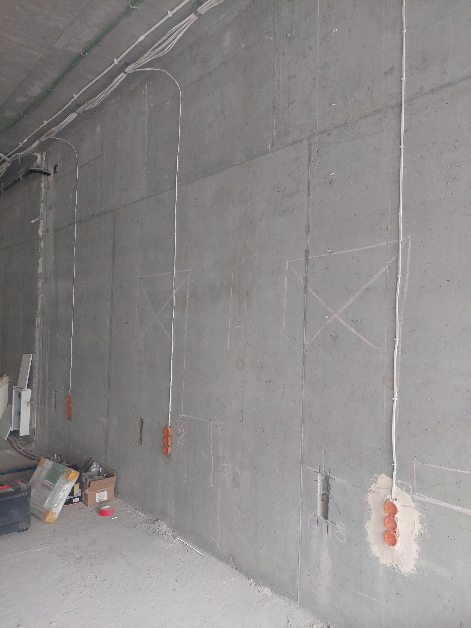 Surowa betonowa ściana z widocznymi przewodami elektrycznymi, puszkami pod gniazdka i zaznaczeniami miejsc instalacji, narzędzia i materiały budowlane na podłodze.