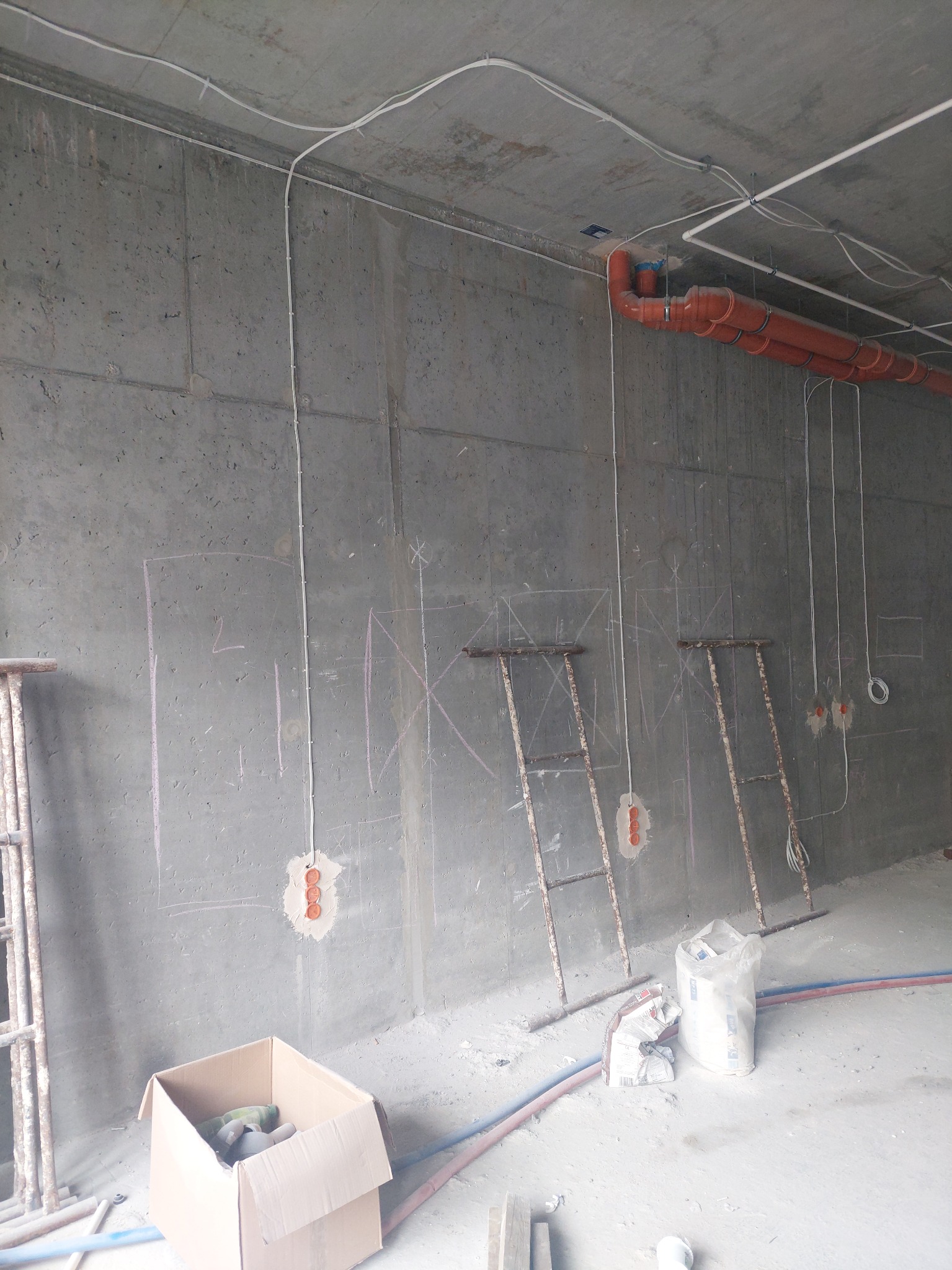 Surowy betonowy pokój w trakcie instalacji elektrycznej: widoczne kable prowadzone po ścianach i suficie, pomarańczowe rury, zaznaczone miejsca na puszki elektryczne, metalowe drabiny, karton...