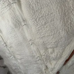 Poddasze pokryte grubą warstwą białej pianki izolacyjnej, przymocowanej metalowymi zszywkami do drewnianych belek stropowych, widoczne fragmenty muru z cegieł.