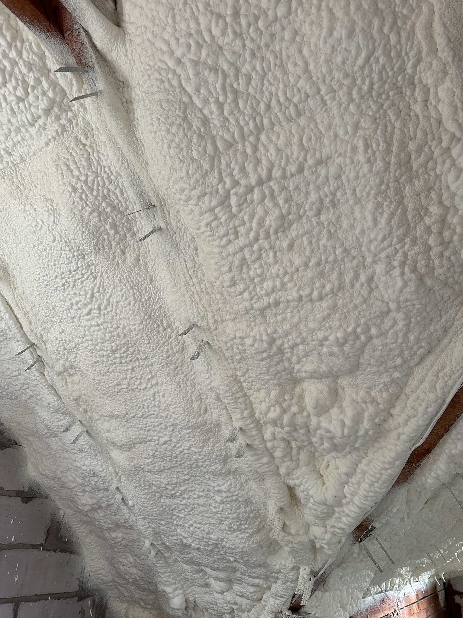 Poddasze pokryte grubą warstwą białej pianki izolacyjnej, przymocowanej metalowymi zszywkami do drewnianych belek stropowych, widoczne fragmenty muru z cegieł.