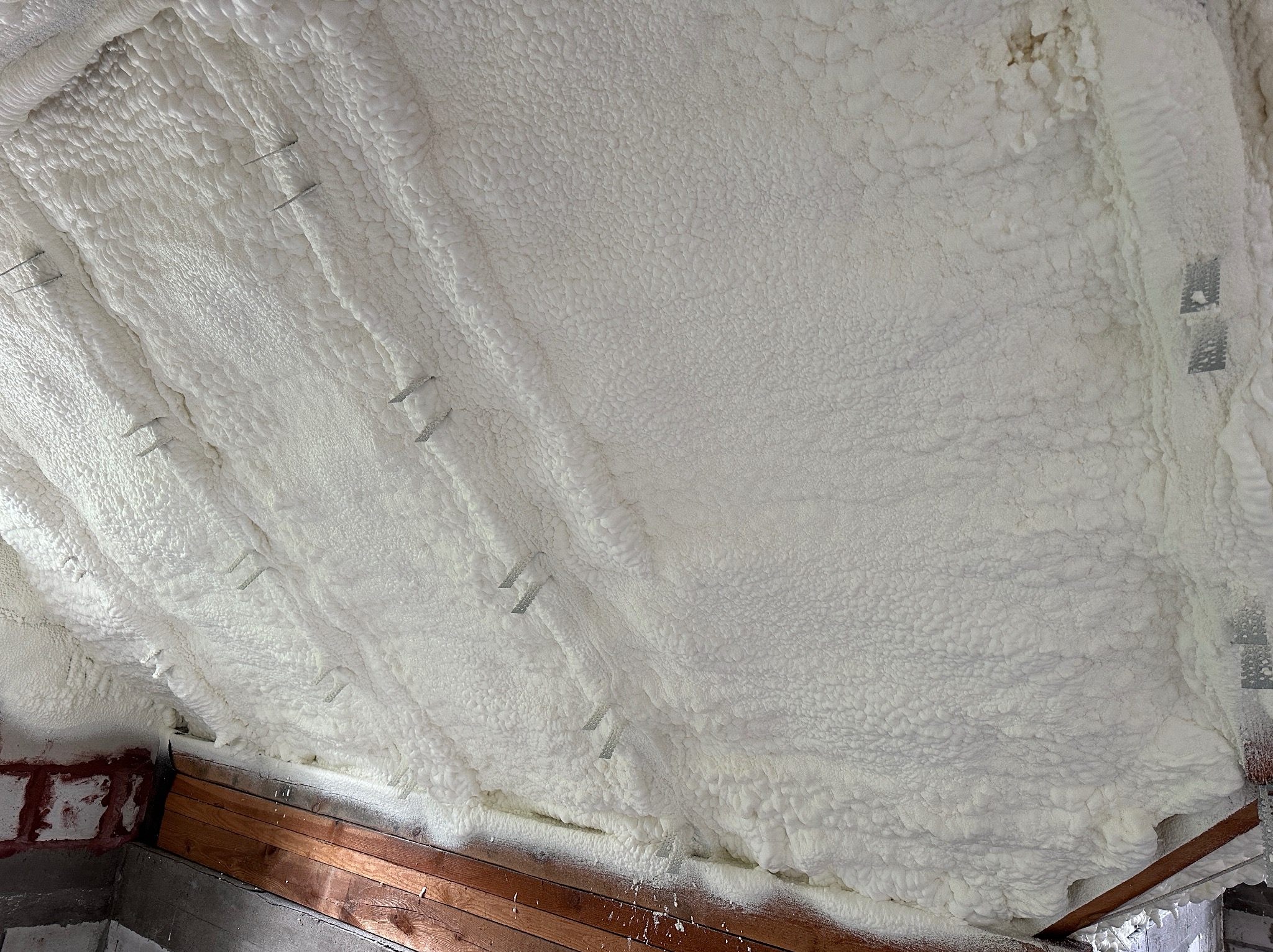 Poddasze z widoczną warstwą białej pianki izolacyjnej, przymocowanej metalowymi kotwami, z fragmentem drewnianej konstrukcji i muru z cegieł w dolnej części kadru.