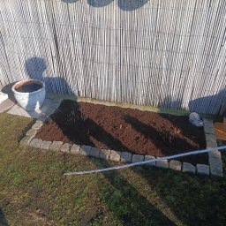 Filip Lubas - Obrzeże rabaty z kostki granitowej, wypełnionej świeżą ziemią, z widocznymi sadzonkami. W tle bambusowy płot i dekoracje ogrodowe.