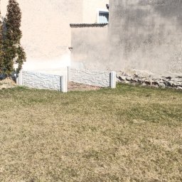 Filip Lubas - Niski murek z prefabrykatów betonowych o fakturze kamienia, oddzielający trawnik od ściany budynku. Widoczne elementy konstrukcyjne i trawa.