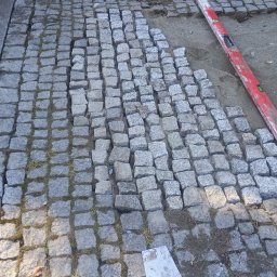 Filip Lubas - Fragment układanej kostki brukowej z granitu. Widoczny poziomica, szpachelka i stos kostki. Częściowo ułożona nawierzchnia z widocznymi spoinami.