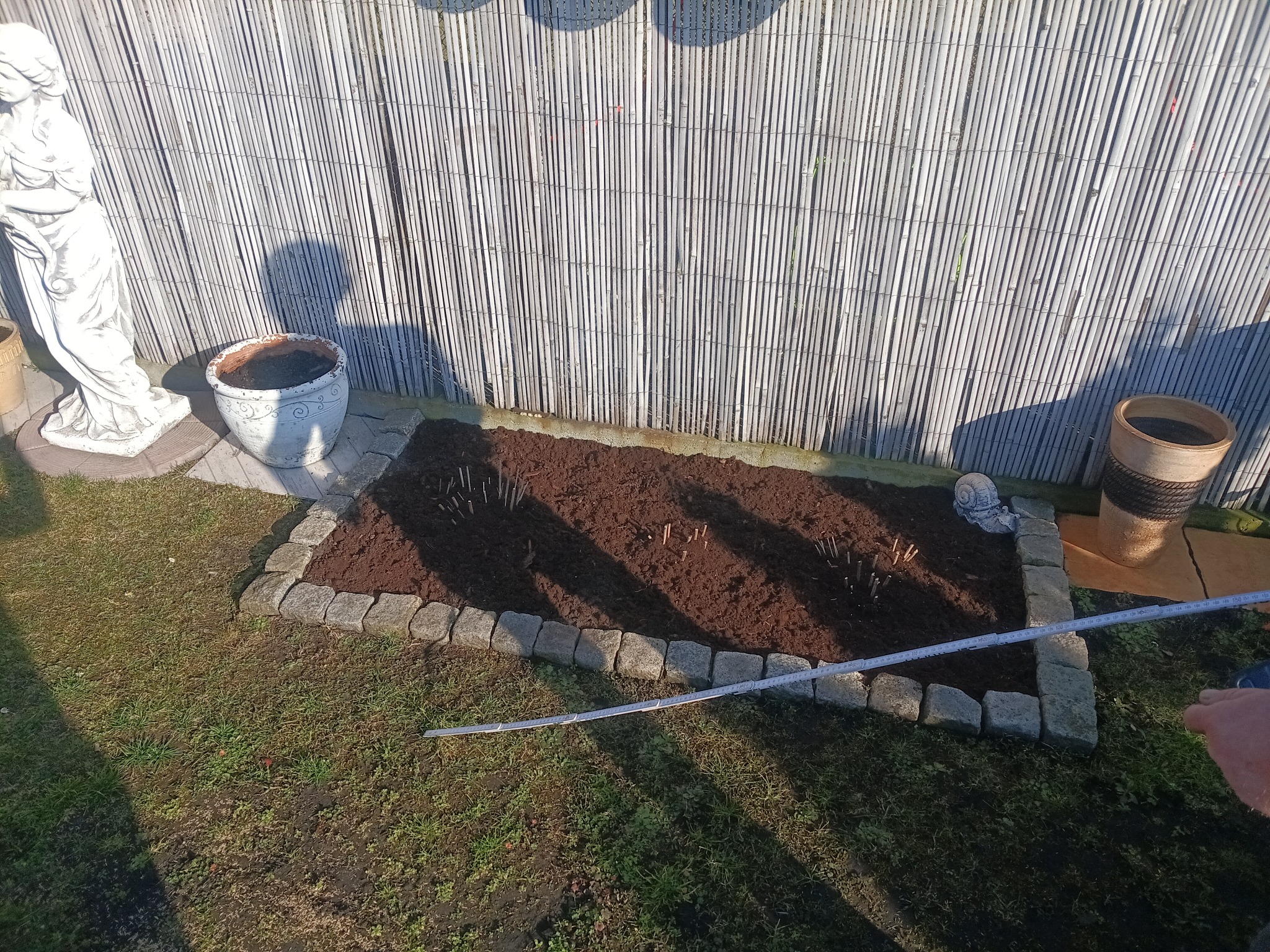 Obrzeże rabaty z kostki granitowej, wypełnionej świeżą ziemią, z widocznymi sadzonkami. W tle bambusowy płot i dekoracje ogrodowe.