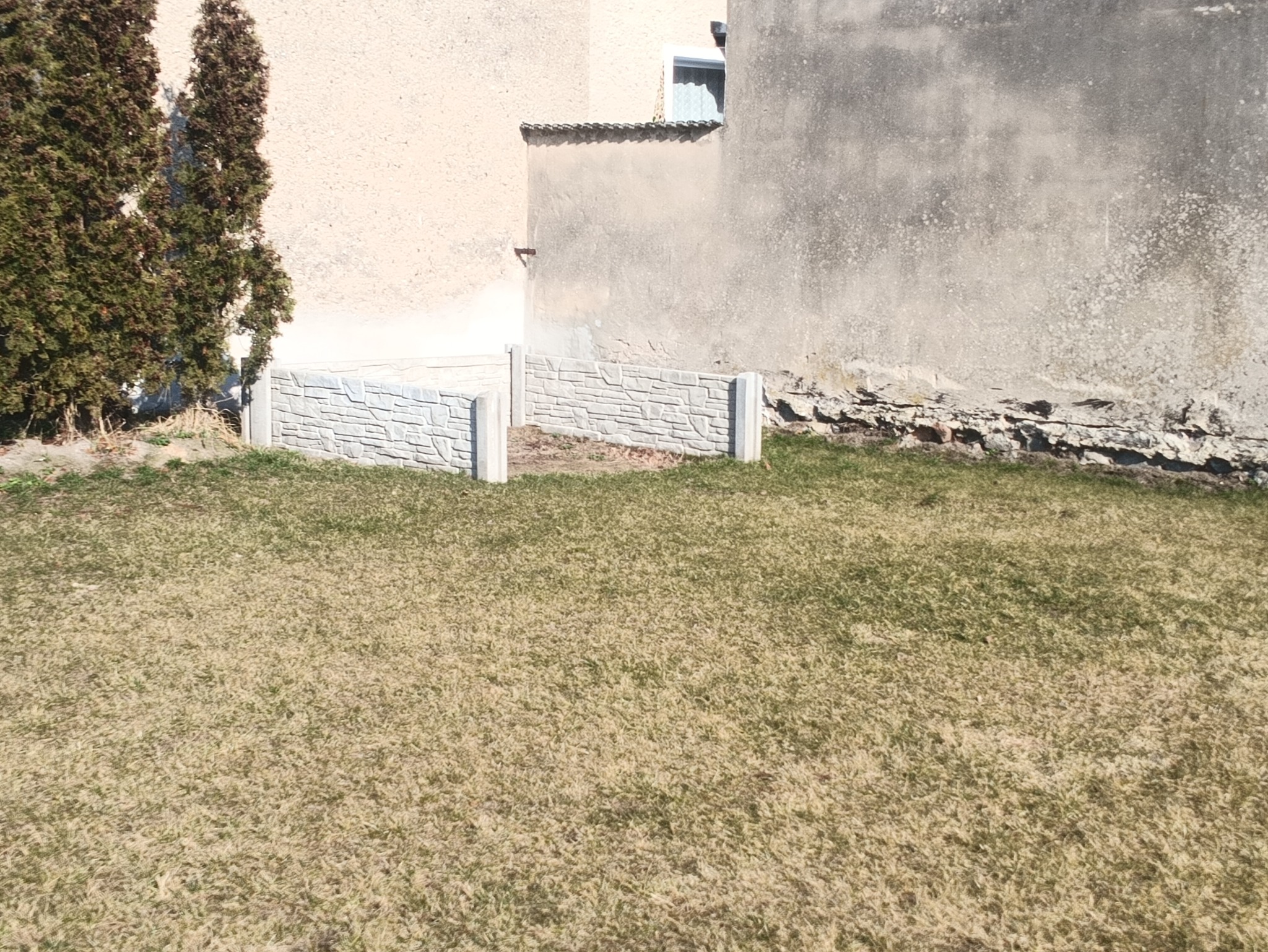 Niski murek z prefabrykatów betonowych o fakturze kamienia, oddzielający trawnik od ściany budynku. Widoczne elementy konstrukcyjne i trawa.