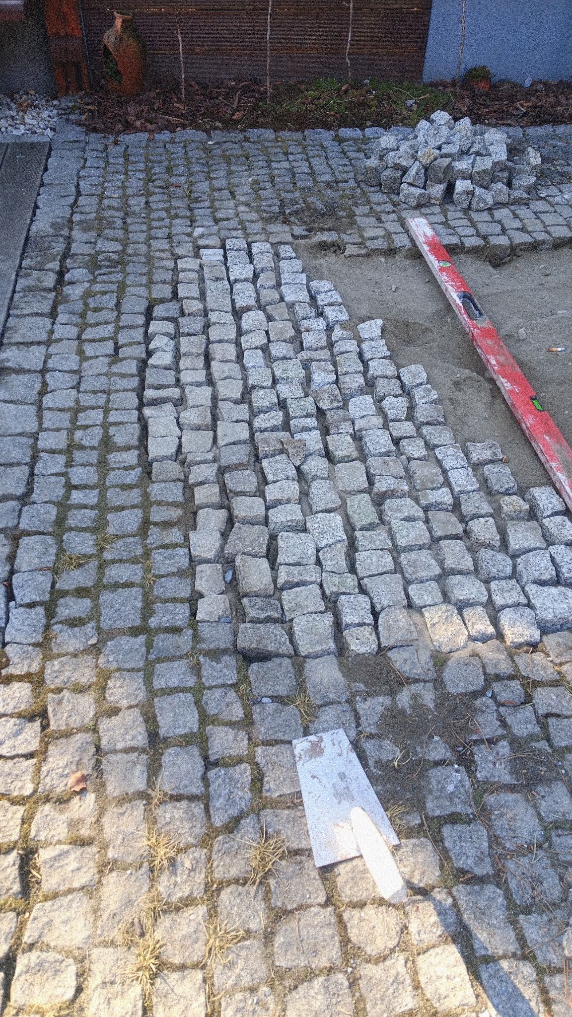 Fragment układanej kostki brukowej z granitu. Widoczny poziomica, szpachelka i stos kostki. Częściowo ułożona nawierzchnia z widocznymi spoinami.
