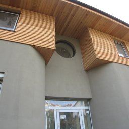 BigRem Plus - Elewacja budynku z szarą, gładką ścianą i drewnianymi panelami ułożonymi poziomo wokół okien oraz okrągłym oknem nad wejściem.