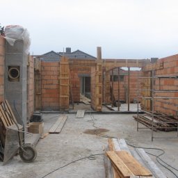 BigRem Plus - Surowy stan budowy domu z czerwonej cegły, z widocznymi drewnianymi szalunkami, wózkiem budowlanym i stertą desek na placu budowy, przy pochmurnej pogodzie.