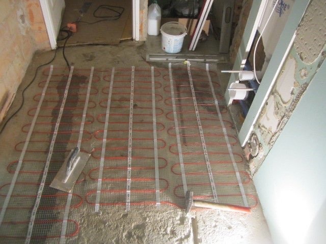 Montaż maty grzewczej na podłodze w łazience, widoczny klej, narzędzia i przygotowane ściany do dalszych prac wykończeniowych.