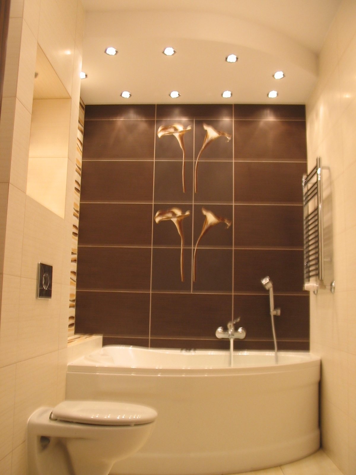 Jasna łazienka z brązowymi płytkami z motywem kwiatowym, narożna wanna, grzejnik drabinkowy i oświetlenie punktowe w suficie.