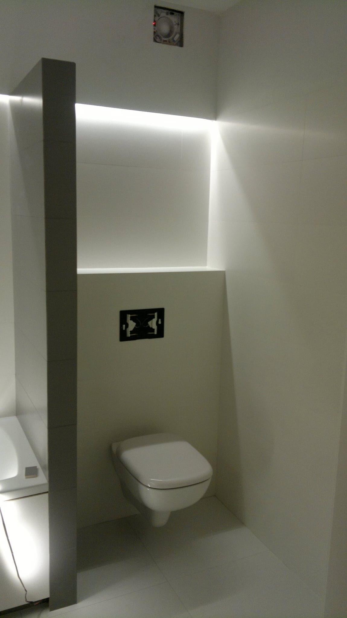 Nowoczesna toaleta podwieszana w minimalistycznej łazience z białymi płytkami, podświetleniem LED ukrytym w suficie i widoczną instalacją wentylacyjną.