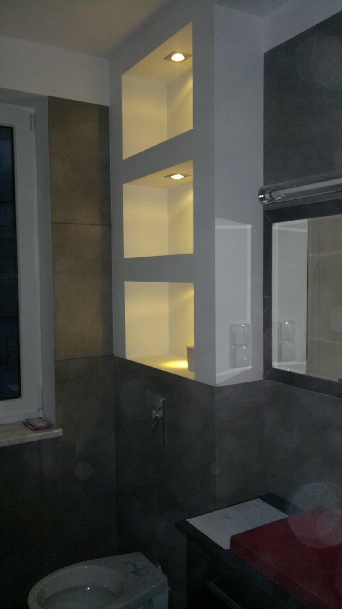 Nowoczesna łazienka z szarymi płytkami, wnękami oświetlonymi punktowo i wiszącą muszlą klozetową.