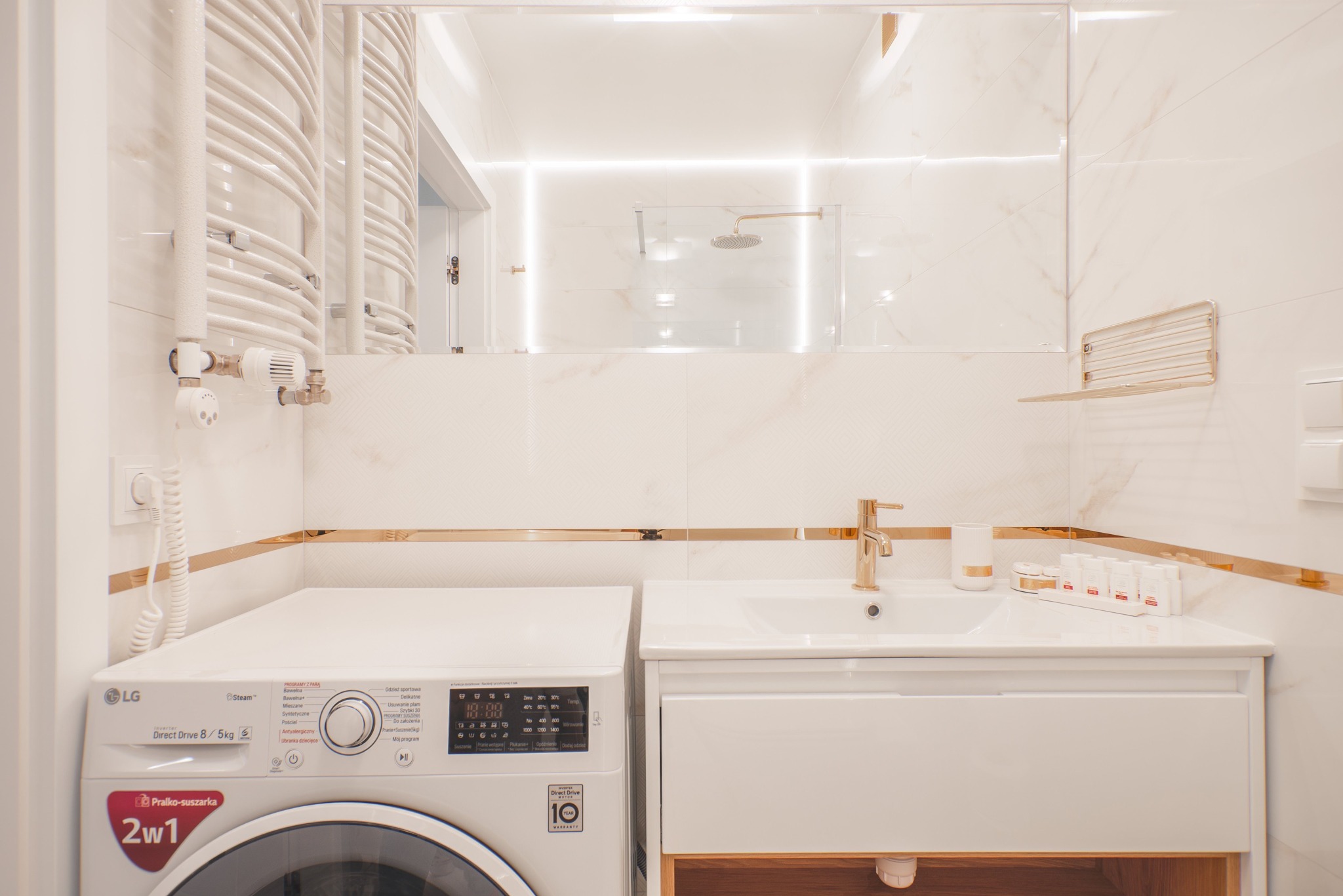 Jasna łazienka z marmurowymi płytkami, złotymi akcentami i pralko-suszarką LG obok umywalki z szafką. W lustrze odbija się kabina prysznicowa. Na blacie umywalki zestaw kosmetyków.