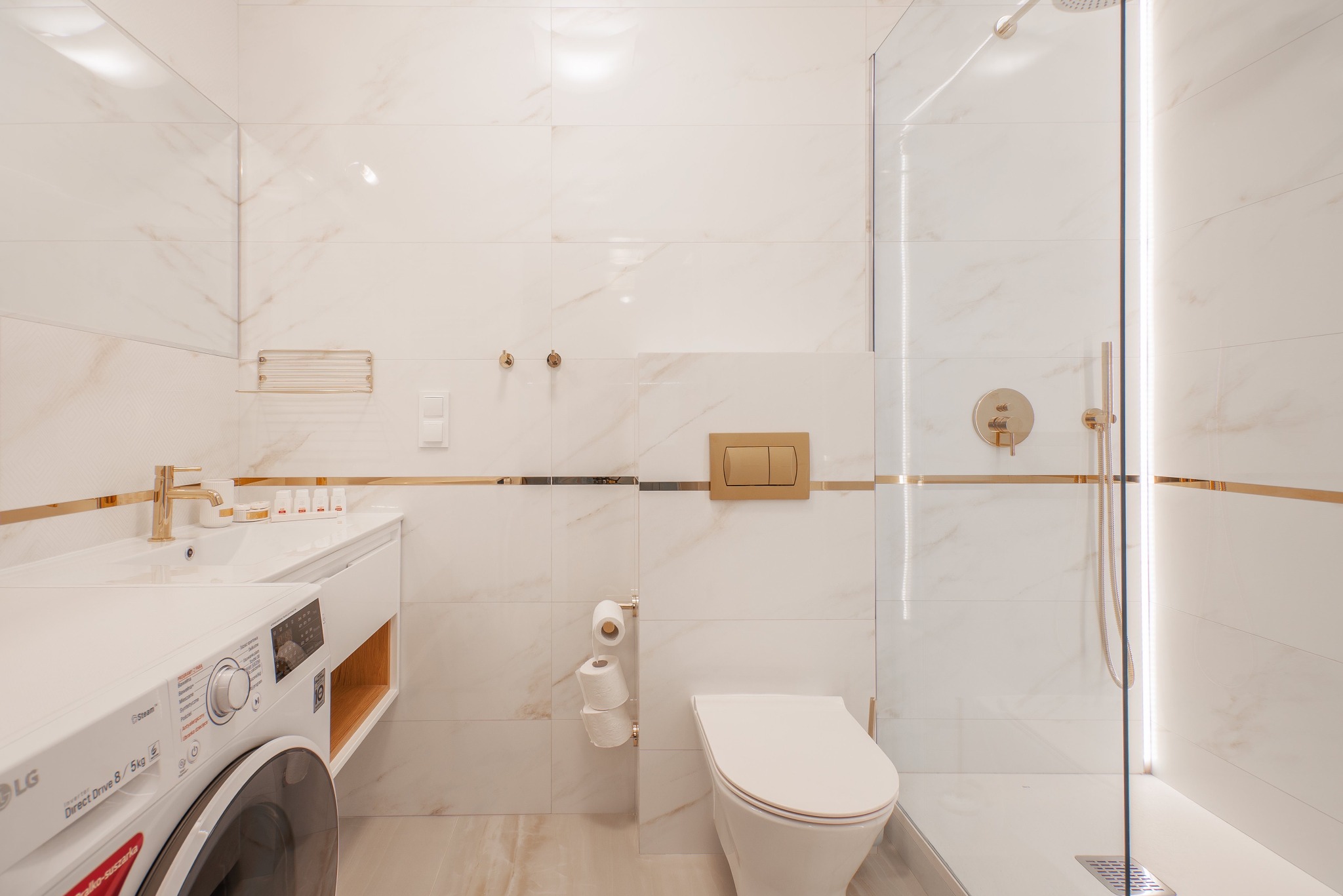 Jasna łazienka z marmurowymi płytkami, złotymi akcentami, kabiną prysznicową ze szklaną ścianką, toaletą, umywalką z szafką i pralką LG.