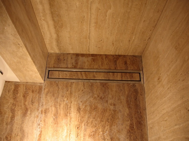 Beżowa ściana i sufit w łazience wykończone trawertynem z zamontowanym liniowym odpływem prysznicowym ze stali nierdzewnej.