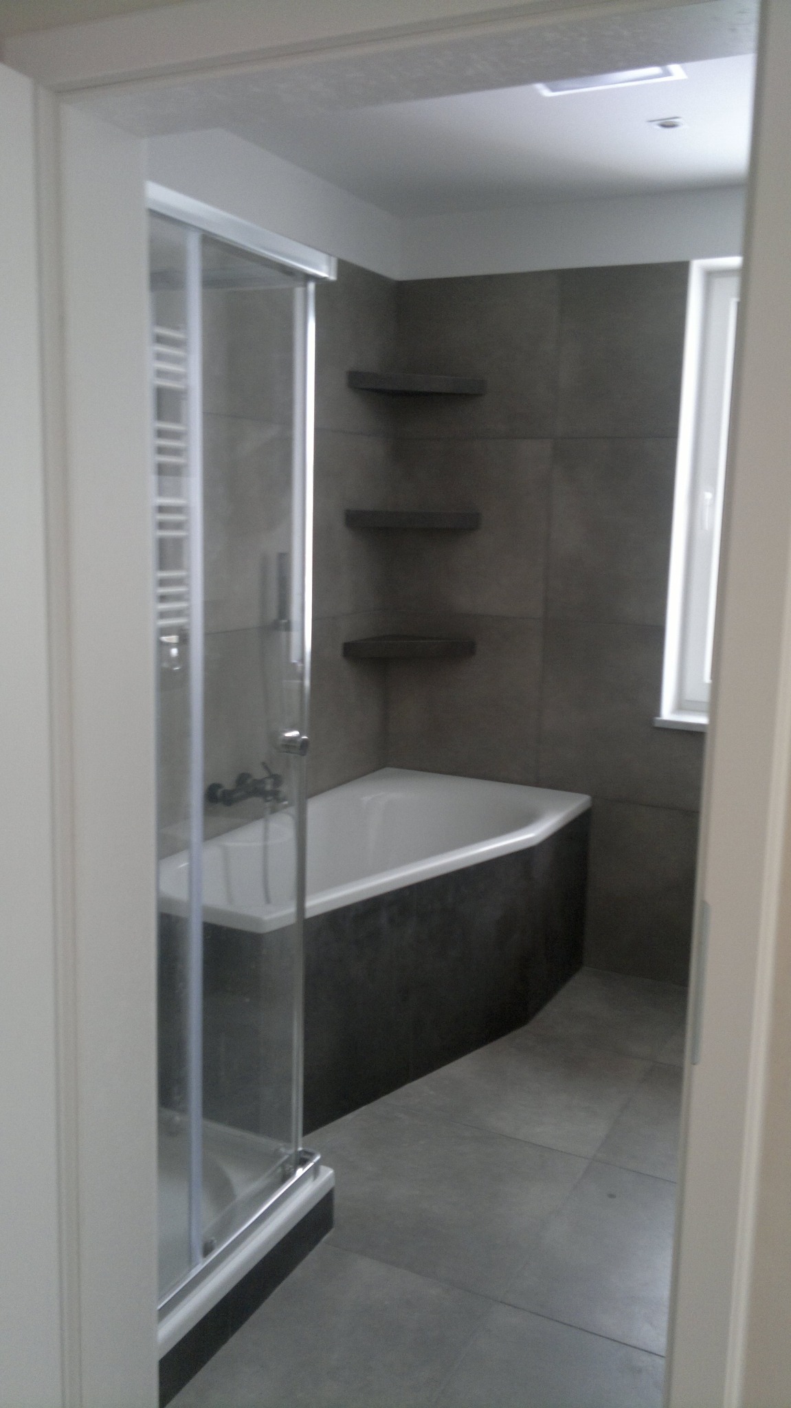 Nowoczesna łazienka z kabiną prysznicową, wanną obudowaną ciemnoszarą płytką, szarymi płytkami na ścianach i podłodze oraz półkami narożnymi.