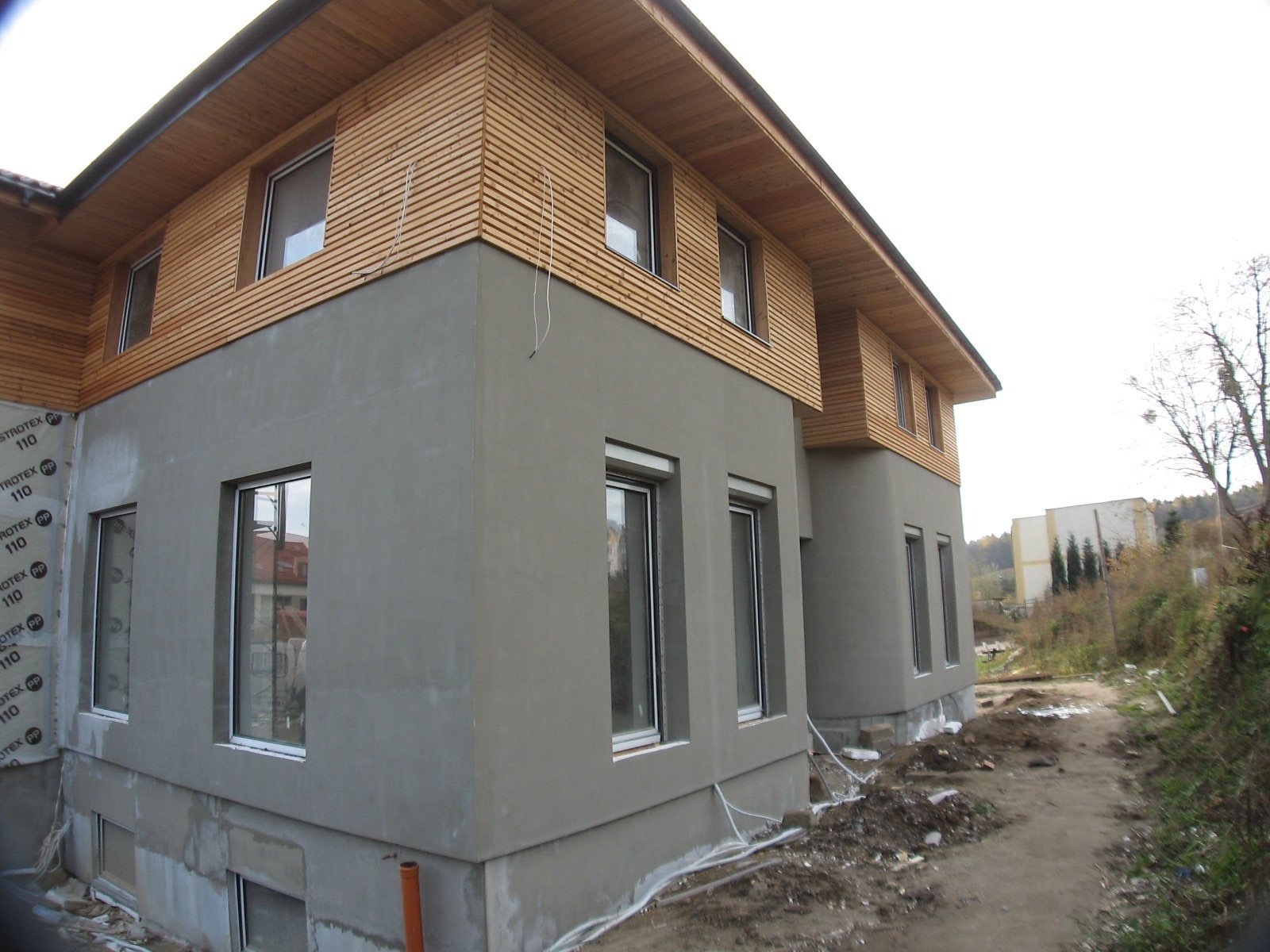 Dwukondygnacyjny budynek w trakcie wykończenia elewacji: dolna część szara, górna wykończona drewnianymi panelami, widoczne okna z roletami i folia ochronna na dolnej części ściany.