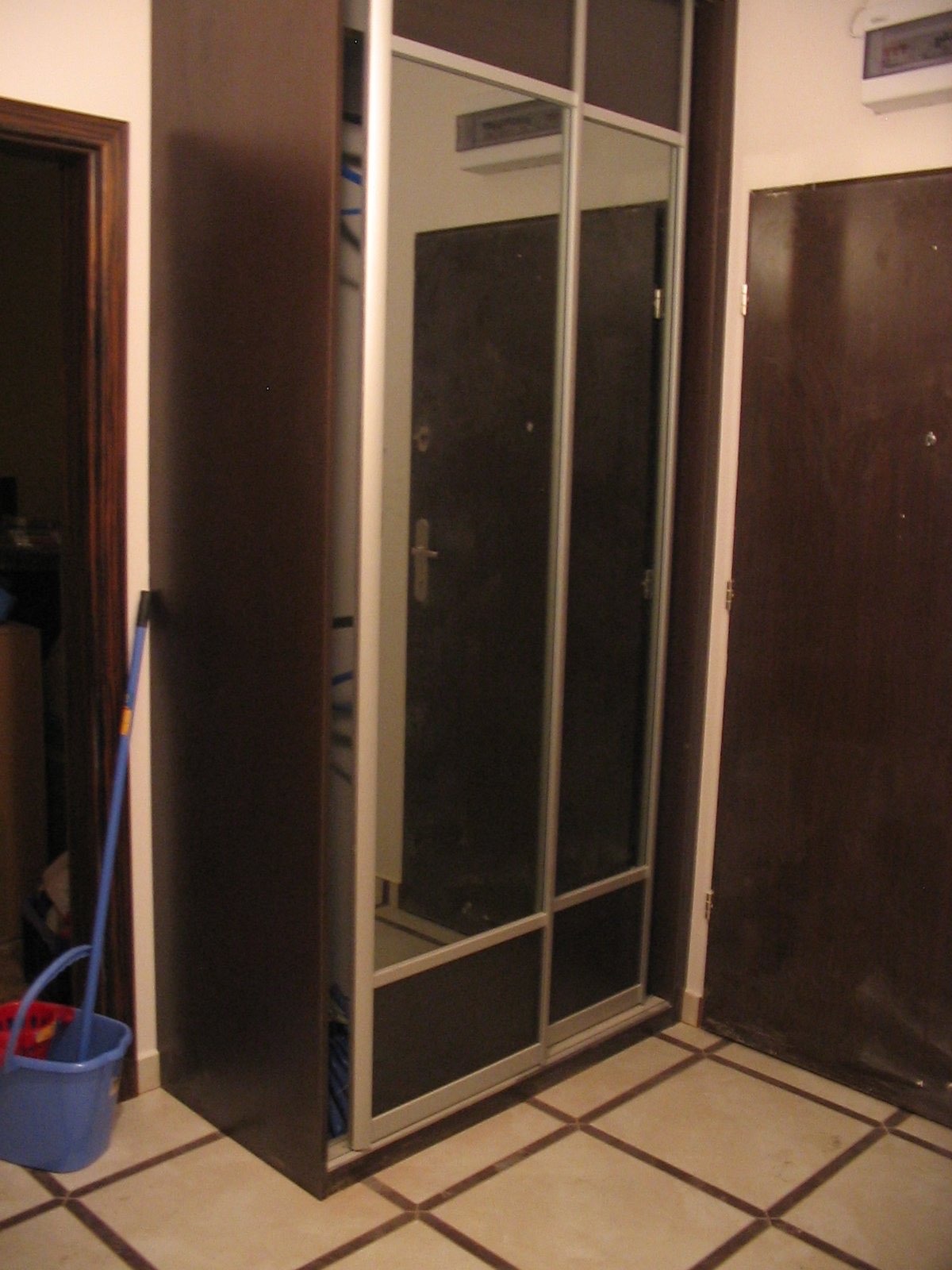 Wnętrze mieszkania z szafą wnękową z przesuwnymi drzwiami z lustrami, widoczny fragment podłogi z kwadratowych płytek oraz drzwi wejściowe.