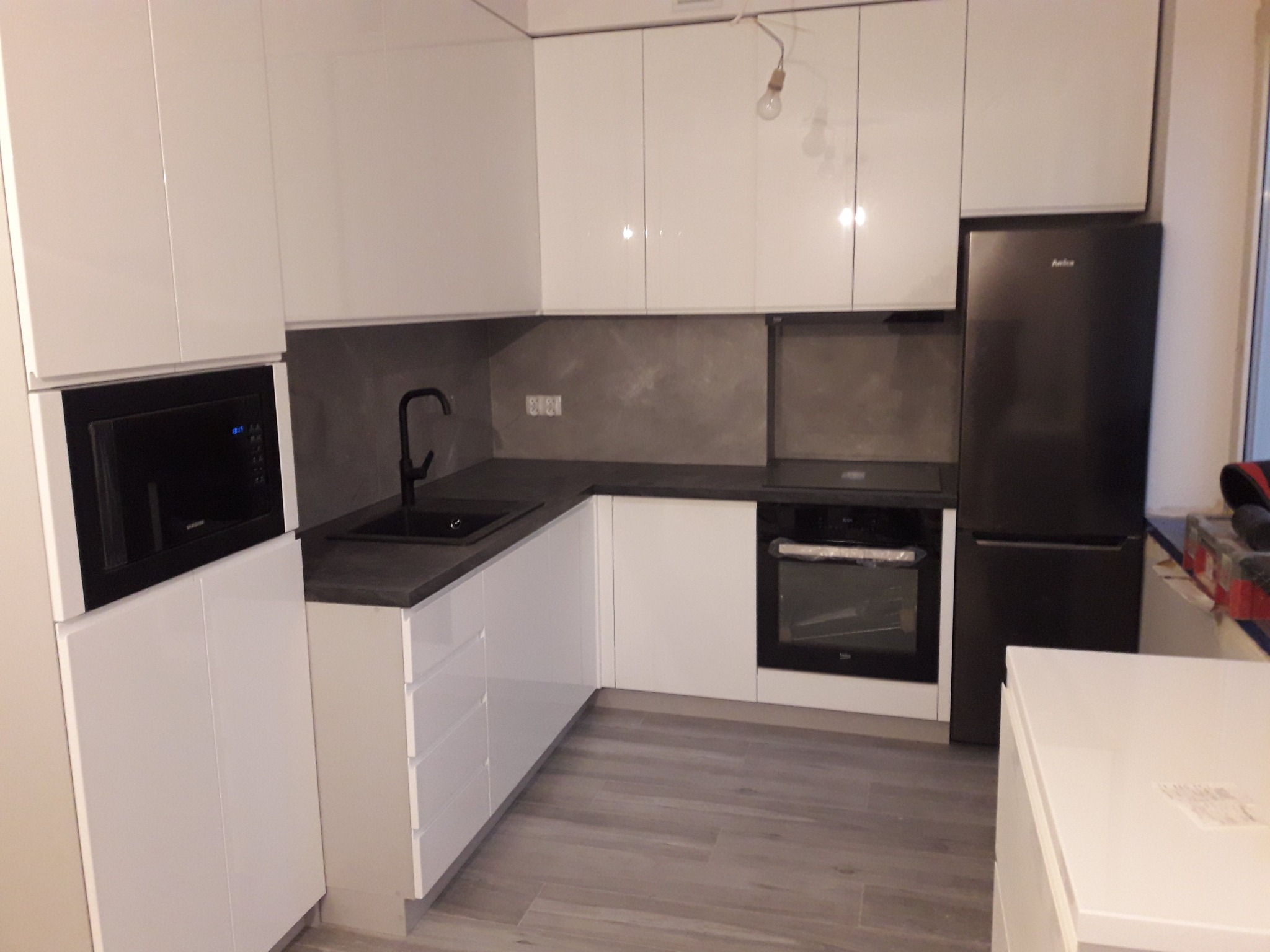 Nowoczesna, biała kuchnia z ciemnym blatem i czarną armaturą, widok na zabudowę kuchenną z piekarnikiem, mikrofalówką i lodówką Amica, surowa ściana nad blatem.