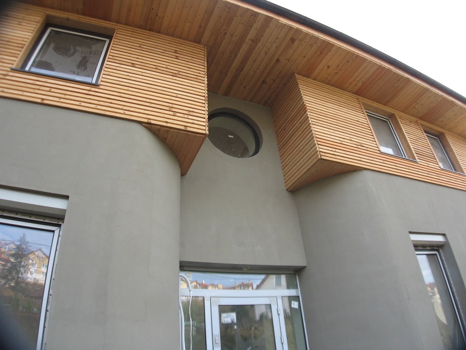 Elewacja budynku z szarą, gładką ścianą i drewnianymi panelami ułożonymi poziomo wokół okien oraz okrągłym oknem nad wejściem.