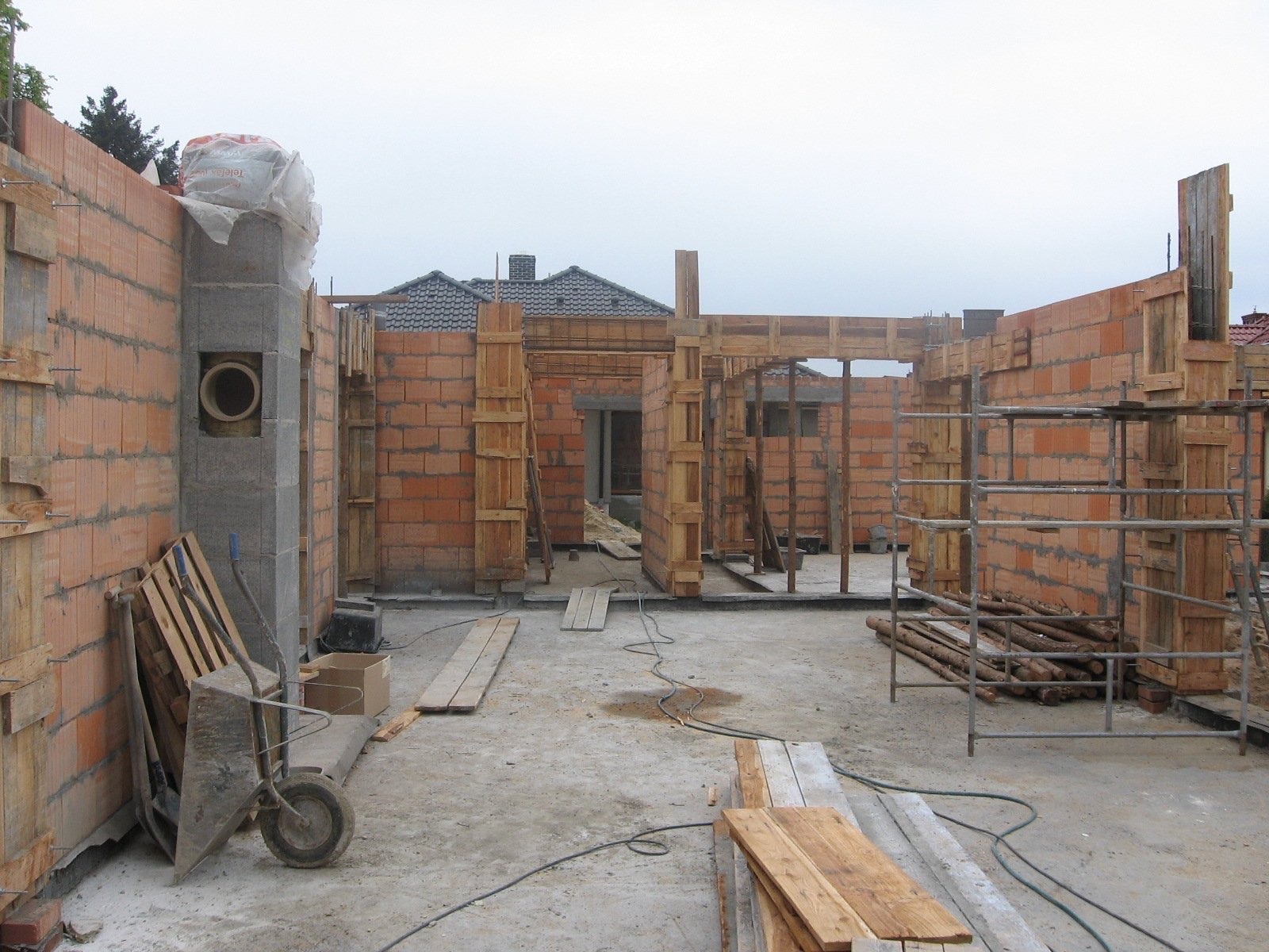 Surowy stan budowy domu z czerwonej cegły, z widocznymi drewnianymi szalunkami, wózkiem budowlanym i stertą desek na placu budowy, przy pochmurnej pogodzie.