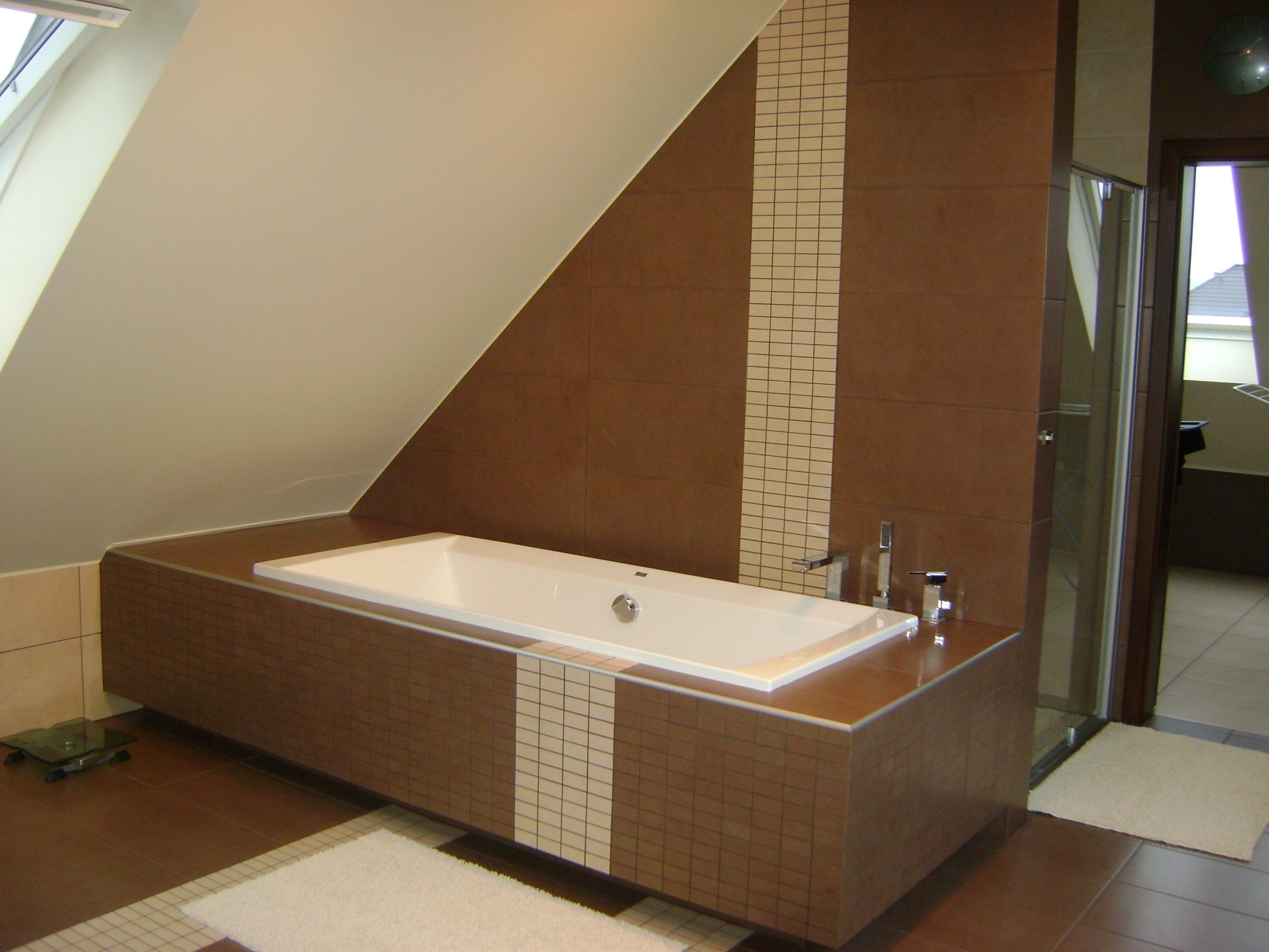 Nowoczesna łazienka na poddaszu z brązowymi płytkami, białą wanną obudowaną płytkami i kabiną prysznicową ze szklanymi drzwiami.