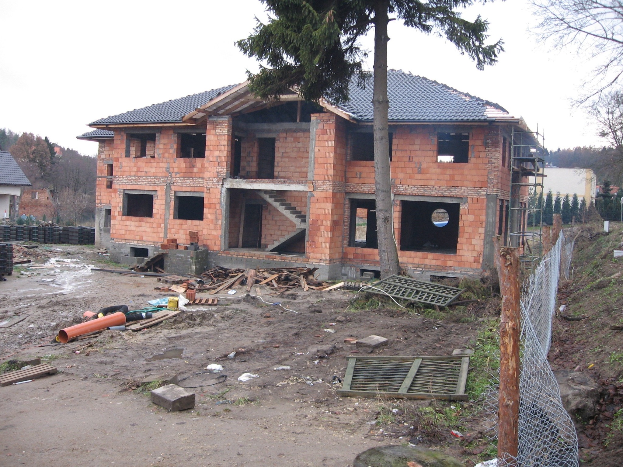 Niedokończony dwupiętrowy dom z cegły z ciemnoszarą dachówką, widoczne schody wewnętrzne, otoczenie błotniste, z porozrzucanymi materiałami budowlanymi i rurami.