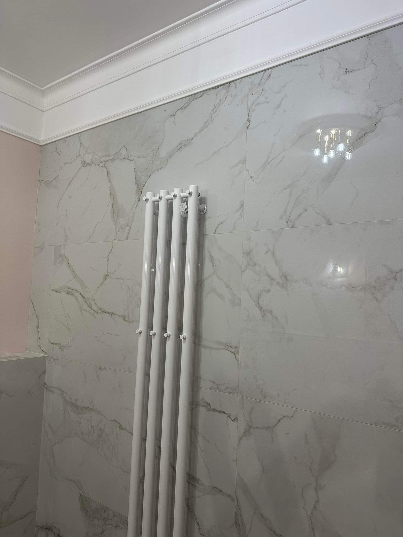Białe płytki imitujące marmur na ścianie łazienki, z widocznym białym grzejnikiem drabinkowym i fragmentem różowej ściany w rogu.