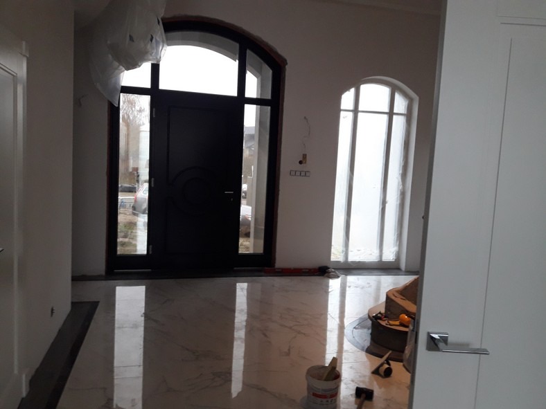 Wnętrze w trakcie remontu z nową, czarną, łukową stolarką drzwiową i okienną, marmurowa posadzka z widocznymi narzędziami glazurniczymi i wiaderkiem po kleju.