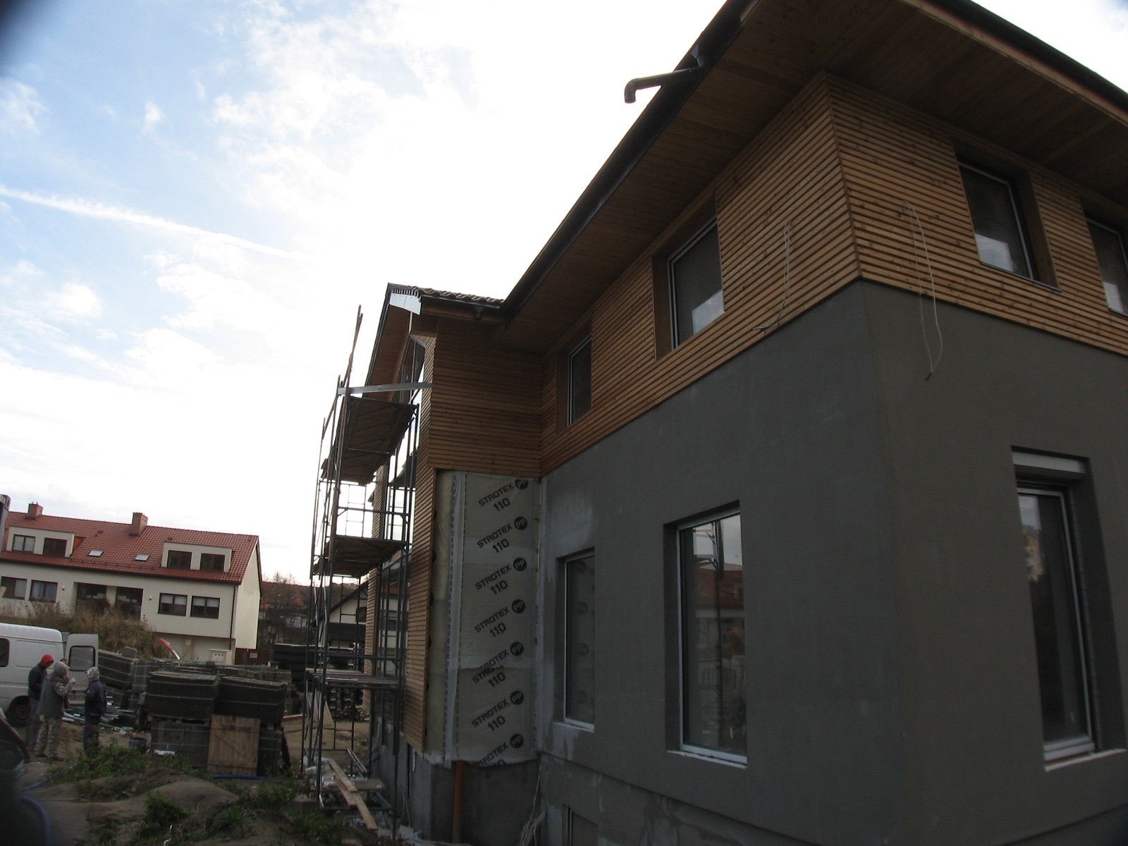 Budynek w trakcie ocieplania i wykańczania elewacji, widoczne rusztowania, ocieplenie styropianem, fragmenty elewacji pokryte drewnem, dolna część elewacji w szarym tynku