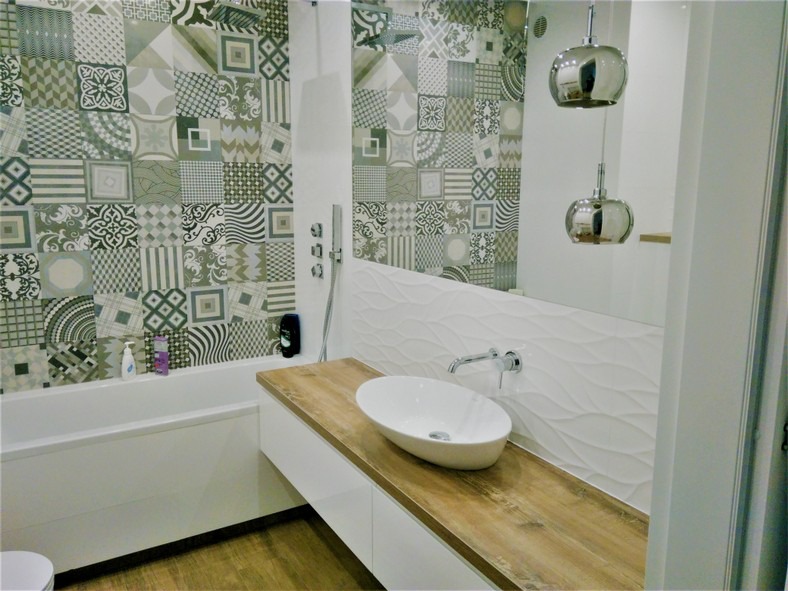 Nowoczesna łazienka z patchworkową ścianą z płytek w odcieniach szarości i zieleni, drewnianym blatem pod umywalką nablatową oraz dekoracyjną, falowaną płytką na ścianie nad blatem. Dwie chromowane...