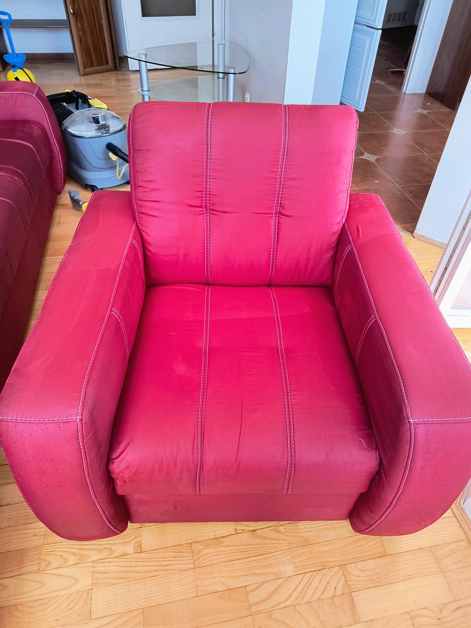 Czerwony fotel tapicerowany z widocznymi śladami użytkowania, obok odkurzacz piorący i fragment drugiego fotela, na drewnianej podłodze.