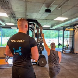 Lifepeak Trainers - Trener personalny obserwuje ćwiczącego klienta w siłowni. Mężczyzna w czarnej koszulce z napisem 'TRENER PERSONALNY' i logo firmy. Ćwiczący wykonuje wykrok z hantlą.