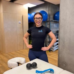 Lifepeak Trainers - Uśmiechnięta kobieta w czarnej koszulce z logo, stojąca w gabinecie z leżanką, piłkami do masażu i taśmami kinesio. W tle widoczne niebieskie piłki.