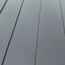 Nowoczesny, grafitowy dach z blachy trapezowej, widok z góry, na tle fragmentu budynku z czarną ramą okienną i zieleni. Widoczne ślady po deszczu na powierzchni.