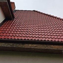 Rafbud - Czerwona dachówka ceramiczna na skośnym dachu domu jednorodzinnego, widok z dołu na tle pochmurnego nieba i fragmentu ściany z oknem.